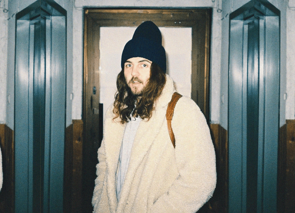  a picture of Hazlett, a twenty-something year old folk guitarist wearing a white, fuzzy coat and a blue knit cap. His long brown hair and beard further express his country-boy appeal. 